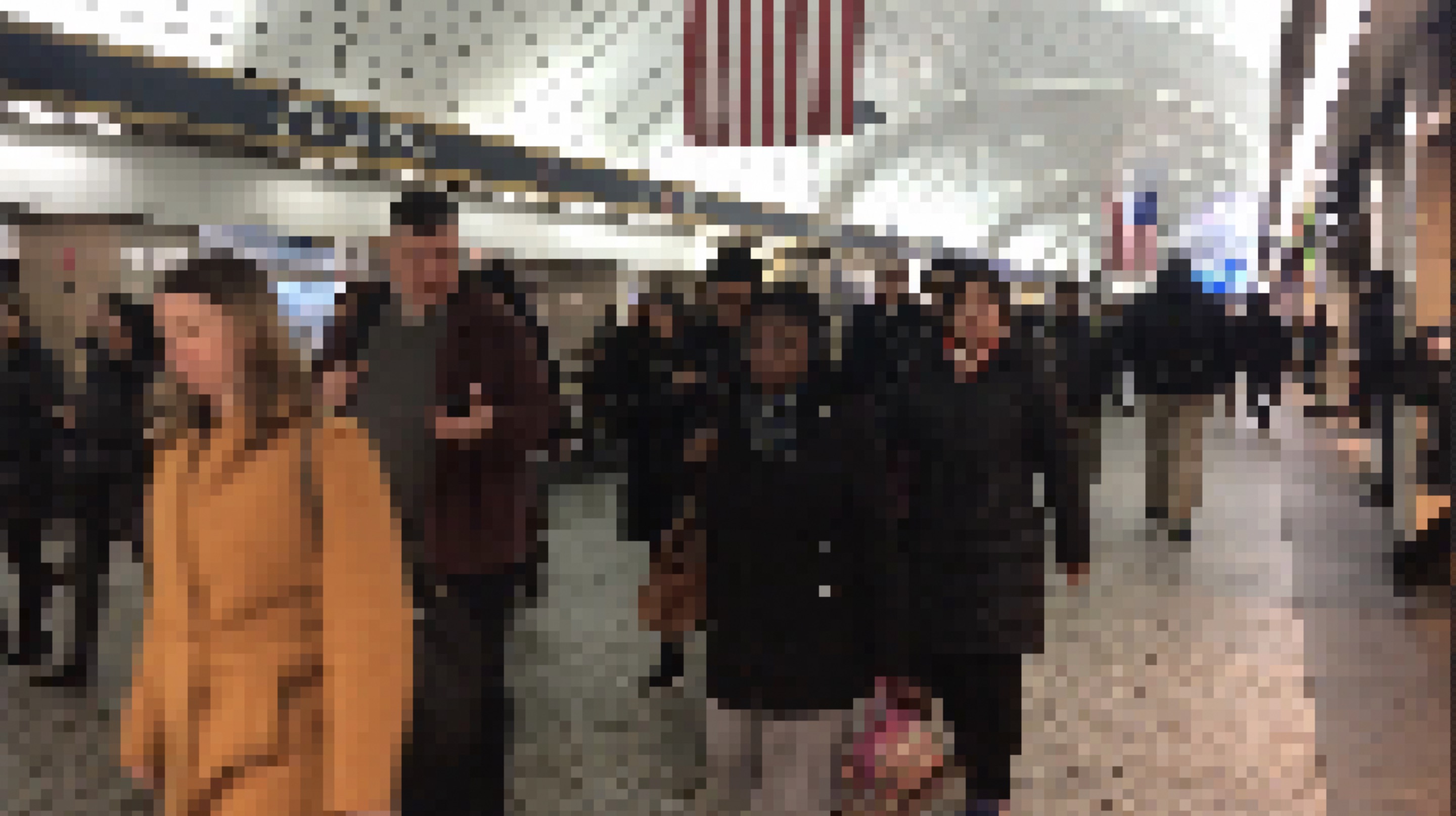 A pixelated photo of people walking through Penn Station train station, the red and white stripes of an American flag hang from the ceiling