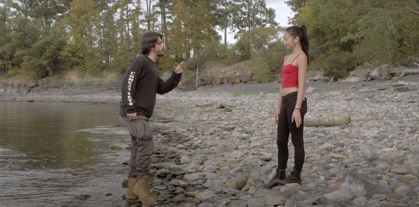 The upper body of a caucasian man with scruffy facial hair and a black shirt pointing a finger at the upper body of an asian woman wearing a red tanktop in an outdoor setting